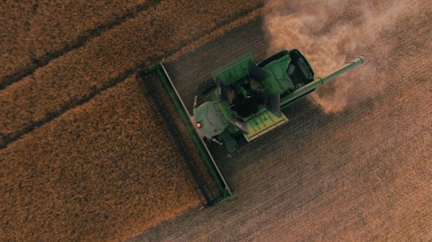 Aerial view of a green combine harvester harvesting a golden field, dust trailing behind it from above.