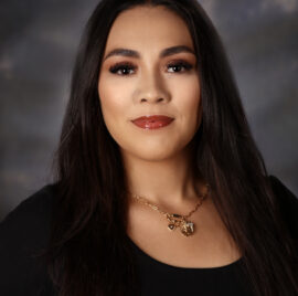 Close-up portrait of a woman with long dark hair, wearing a black top and gold heart necklace, neutral expression against a dark backdrop.