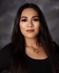 Portrait of a woman with long dark hair, wearing a black top and gold necklace, looking at the camera.
